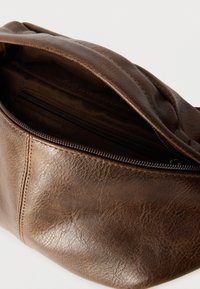 Καφέ δερμάτινη τσάντα με απαλή, υφή επιφάνεια, που διαθέτει φερμουάρ στο επάνω μέρος και ευρύχωρο, χωρίς επένδυση εσωτερικό.