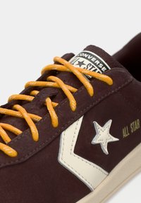 Zapatillas de ante marrón con cordones naranjas, acentos crema, logo de estrella y el texto "All Star" en el lateral, con una superficie texturada.