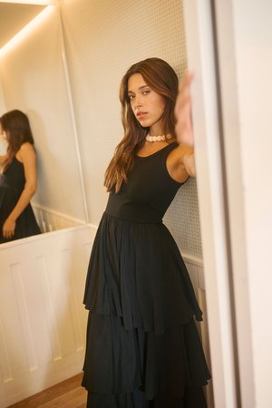 Frau mit langem braunem Haar, die ein ärmelloses schwarzes Stufenkleid und eine massige Halskette trägt, lehnt an einer hellen Wand im Innenraum.