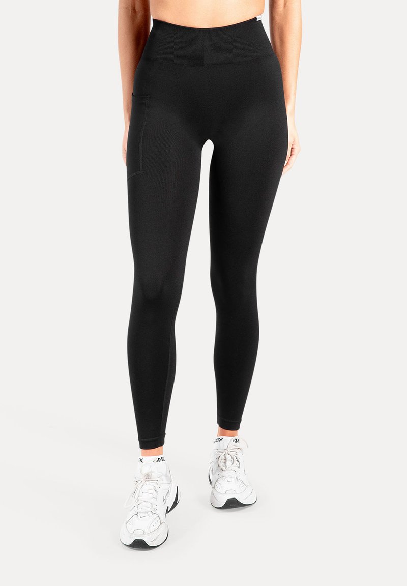 Leggings noirs taille haute en tissu extensible avec une poche latérale. Texture lisse, coupe ajustée, associés à des baskets blanches.