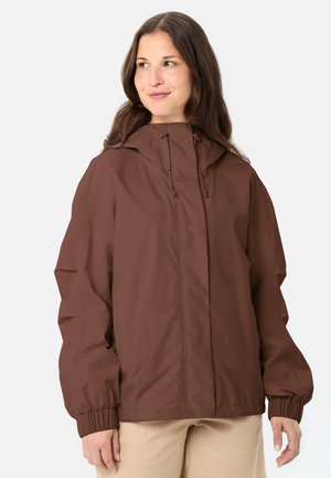 Donna che indossa una giacca marrone con cappuccio e polsini elasticizzati e pantaloni beige, che guarda leggermente a sinistra su uno sfondo neutro.