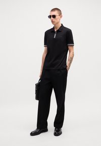 Camisa polo preta com colarinho e detalhes nas mangas cinzentos, combinada com calças pretas e sapatos pretos. Acessórios incluem óculos de sol escuros e uma mala.