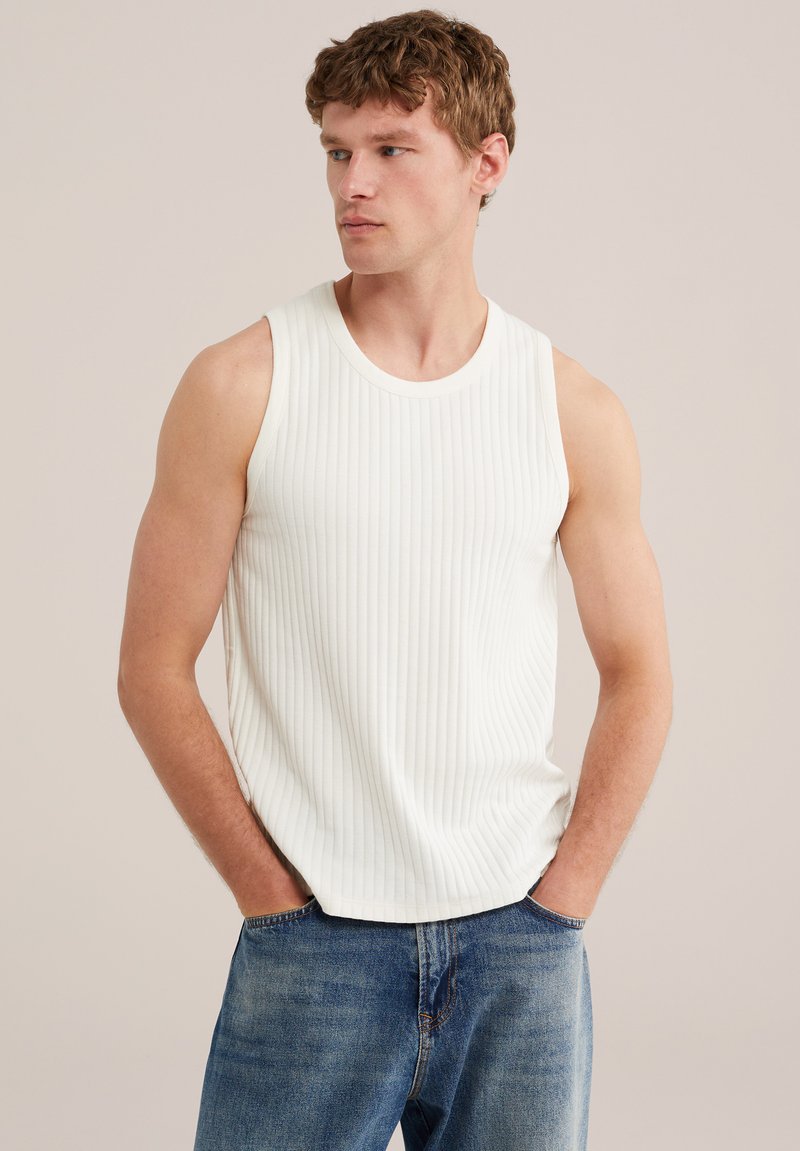 Jeune homme aux cheveux courts et bouclés portant un débardeur blanc côtelé et un jean bleu, les mains dans les poches, regardant sur le côté.
