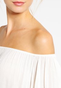 Off-shoulder, vit blus tillverkad av lätt, veckad tyg med elastisk halsringning och mjuk textur, som visar upp en avslappnad passform.