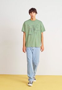 T-shirt in cotone verde con stampa grafica, abbinato a jeans azzurri chiari e scarpe da ginnastica nere e bianche. Sfondo semplice, posa in piedi.