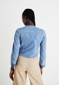 Svetlo modra denim jakna z ožjim krojem, dolgimi rokavi in zaobljenim robom, v kombinaciji s bež hlačami visoke pasu, ki imajo zadnje žepke.