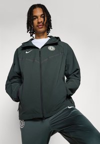 Mørkegrønn Nike-jakke med glidelås, hette og Chelsea FC-logo på brystet. Har grå detaljer og et lett, teksturert stoff.