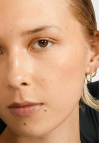 Suure lähedalt on näha naist, kellel on hele nahk, pruuni silmad, lühikesed blondid juuksed ja paremas kõrvas kaks kuldset ringi, millel on bambuse muster.