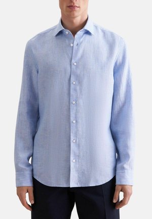 Mand iført en lyseblå, langærmet skjorte med knapper og krave samt sorte bukser, stående foran en ensfarvet baggrund.