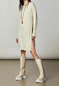 Vestido de punto beige claro con mangas largas y aberturas laterales. Combinado con botas crema hasta la rodilla que tienen una suela gruesa blanca y cremallera lateral.