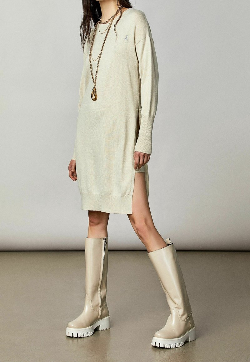 Vestido de punto beige claro con mangas largas y aberturas laterales. Combinado con botas crema hasta la rodilla que tienen una suela gruesa blanca y cremallera lateral.