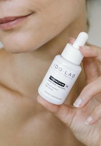 Biała szklana butelka z kroplomierzem oznaczona napisem "IDO LAB TROXACALM" z czarnym tekstem, zawierająca serum do twarzy kontrolujące zaczerwienienia, trzymana w dłoni.