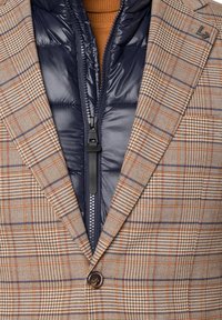 Brązowa marynarka w kratę z wyłogowym kołnierzem, pojedynczym zapięciem na guzik oraz kontrastową kamizelką puchową w granatowym kolorze z zamkiem błyskawicznym z przodu.