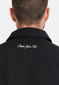 Hombre con cabello corto y aretes de aro dorados, vestido con una camiseta negra de canalé con "Sixth June, Co." bordado en el cuello de la parte posterior.