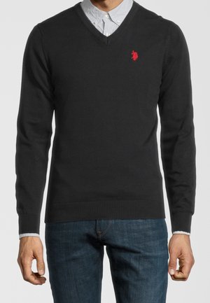 Mann trägt schwarzen V-Ausschnitt-Pullover mit rotem Logo über hellblauem Hemd und blauer Jeans, steht vor einem einfarbigen Hintergrund.