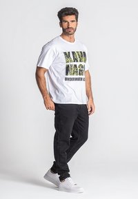 Bijela majica s crnim i zelenim grafičkim tekstom, u kombinaciji s crnim hlačama i bijelim tenisicama. Model stoji na neutralnoj pozadini.