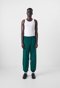 Calças verdes, leves e soltas, com cintura elástica e punhos nos tornozelos. Apresenta um pequeno rótulo roxo na lateral. Top de alças branco.