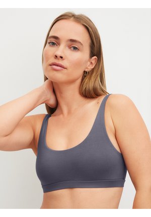 Vrouw met blond haar, gekleed in een grijze mouwloze cropped top, poseert met één hand achter haar nek tegen een effen achtergrond.