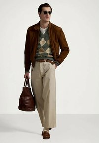 Casaco de camurça castanho sobre um suéter verde com padrão argyle, calças largas bege, mocassins castanhos e uma mala de couro castanha. Óculos de sol redondos completam o visual.