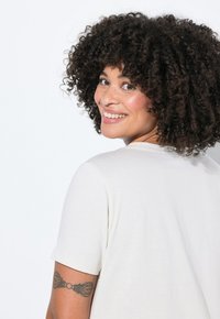 Camiseta de color beige claro con un corte relajado y mangas cortas. Textura suave, diseño de cuello redondo y tatuaje visible en el brazo.
