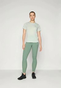 Junge Frau steht in einem hellgrauen T-Shirt, salbeigrünen Leggings und schwarzen Sportschuhen vor einem schlichten hellen Hintergrund.