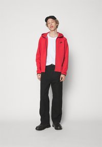 Felpa rossa con zip e logo nero, abbinata a pantaloni neri a gamba larga e stivali neri. Sotto, una semplice t-shirt bianca.