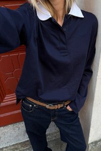 Marineblaues Langarm-Polohemd mit weißem Kragen, ausgestattet mit einer Knopfleiste. Getragen mit dunklen Denim-Jeans und einem braunen Gürtel.