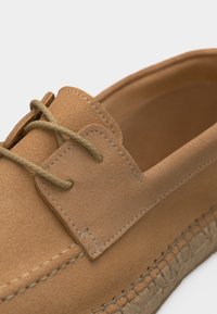 Mocassin en daim beige avec lacets tressés et couture détaillée le long de la tige et de la semelle, mettant en valeur un savoir-faire doux et un style décontracté.