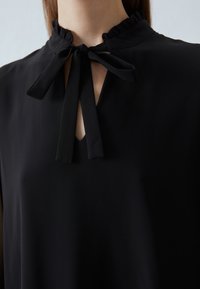Blusa nera con colletto a volants e apertura a chiave sul davanti. Presenta un nastro a fiocco e un tessuto liscio e leggero.
