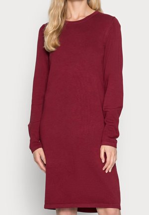 Vrouw in een langmouw, knie-lange, effen kastanjebruine sweaterjurk met een ronde halslijn, tegen een effen achtergrond.