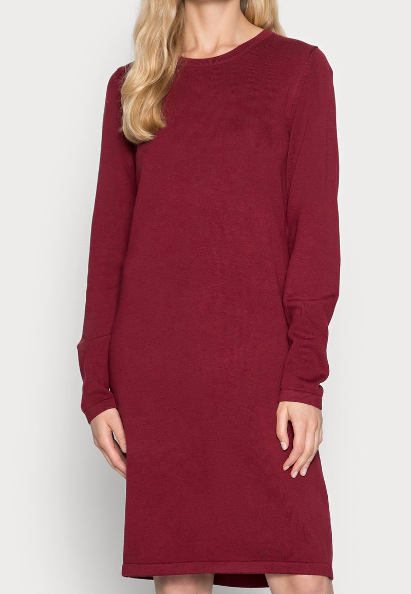 Vrouw in een langmouw, knie-lange, effen kastanjebruine sweaterjurk met een ronde halslijn, tegen een effen achtergrond.