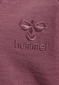 Un textil color marrón con un logo bordado de una abeja y la palabra "hummel", con una textura suave y lisa y un patrón de tejido uniforme.