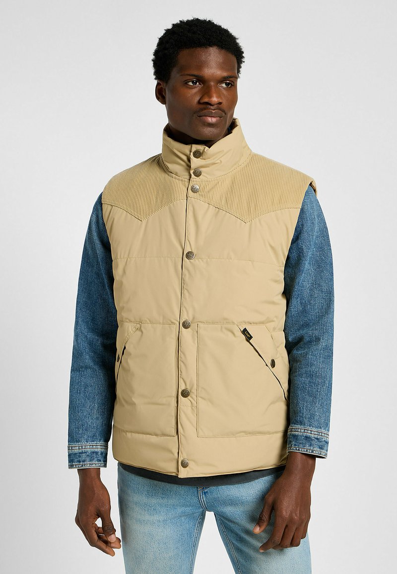 Tannad väst med kordflöjelsaccent på axlarna och denimärmar. Har tryckknappar och två framfickor. Slät textur över hela.