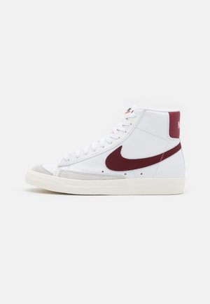Basket blanche montante avec logo Nike bordeaux et patch au talon, lacets blancs et semelle blanc cassé, vue de côté sur fond uni.