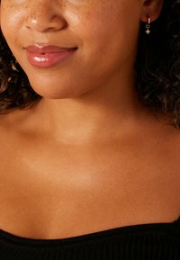 Boucle d'oreille créole en or avec un petit pendentif en forme d'étoile suspendue à l'oreille. Le teint de la peau est chaud avec des taches de rousseur visibles. L'arrière-plan et les détails du corps sont minimalistes.