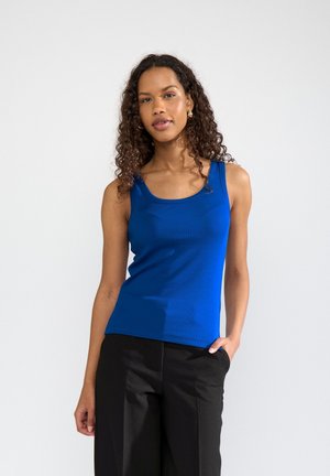 Femme aux cheveux bouclés portant un haut sans manches bleu et un pantalon noir, posant avec une main dans la poche sur un fond uni.