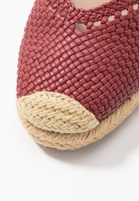 Gewebtes burgunderfarbenes Lederoberteil mit Ausschnitten und einer Zehenkappe aus Juteseil. Die hellbraune gewebte Unterseite sorgt für Kontrast und Textur.