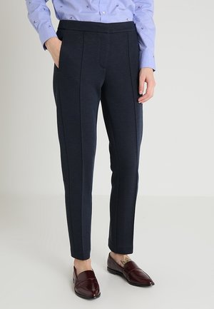Personne portant un pantalon noir ajusté avec des coutures à l'avant, une chemise bleu clair boutonnée, et des mocassins en cuir rouge foncé, debout avec une main dans la poche.