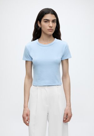 Νέα γυναίκα με μακριά σκούρα μαλλιά φοράει ανοιχτό μπλε εφαρμοστή μπλούζα με κοντά μανίκια και λευκό ψηλόμεσο παντελόνι σε μονόχρωμο φόντο.