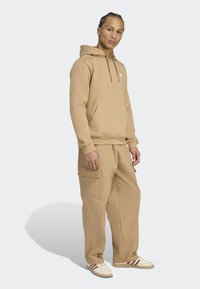 Joven con cabello trenzado que lleva una sudadera color beige, pantalones cargo anchos y zapatillas blancas con rayas marrones, de pie frente a un fondo claro.