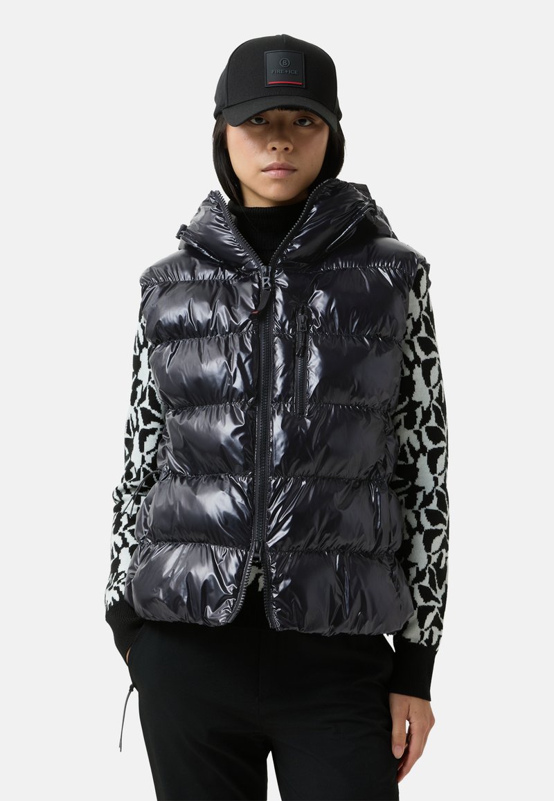 Puszysta czarna kamizelka o błyszczącej, pikowanej teksturze, z zapięciem na zamek i przymocowanym kapturem, nałożona na sweter w wzory i czarne spodnie.