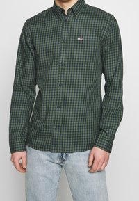 Camisa de cuadros verde y azul marino con botones y cuello, con un pequeño logo y un bolsillo en el pecho. Hecha de tela ligera.