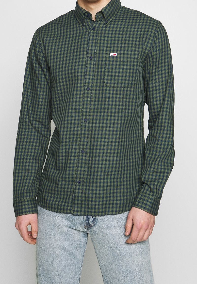 Camisa de cuadros verde y azul marino con botones y cuello, con un pequeño logo y un bolsillo en el pecho. Hecha de tela ligera.