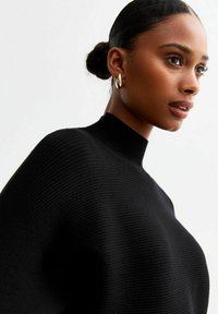 Pull en tricot noir avec un col montant, offrant une texture côtelée. Le modèle porte des boucles d'oreilles en forme de goutte dorée sur un fond neutre.