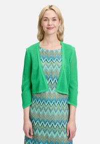 Grüne gestrickte Strickjacke mit Dreiviertelärmeln und offenem Vorderteil, über einem mehrfarbigen Zickzackmuster-Kleid getragen.