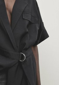 Nahaufnahme einer Person, die eine schwarze, kurzärmelige Jacke mit Gürtel, breitem Revers und aufgesetzter Brusttasche auf der linken Seite trägt.