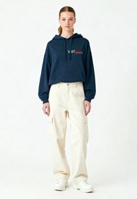 Felpa blu navy con testo ricamato rosso e tasca frontale, abbinata a pantaloni cargo crema con due tasche laterali e sneakers bianche.