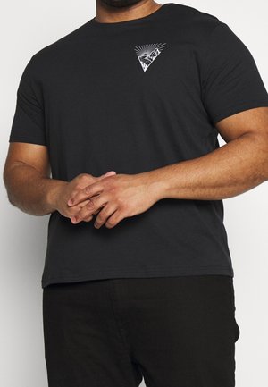 Mand iført sort t-shirt med korte ærmer med lille hvid grafik af bjerg og sol på venstre bryst, hænder løst foldet foran.