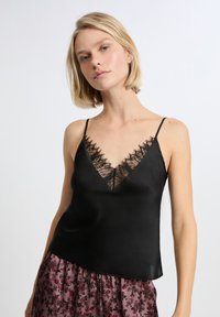 Camisole in satin nero caratterizzata da un profondo scollo a V decorato con delicati bordi in pizzo nero e sottili spalline regolabili.