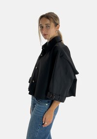 Schwarze, kurz geschnittene Jacke mit hohem Kragen, kontrastierenden Knöpfen und weiten Ärmeln mit Schnallendetails. Getragen über blauen Jeans.